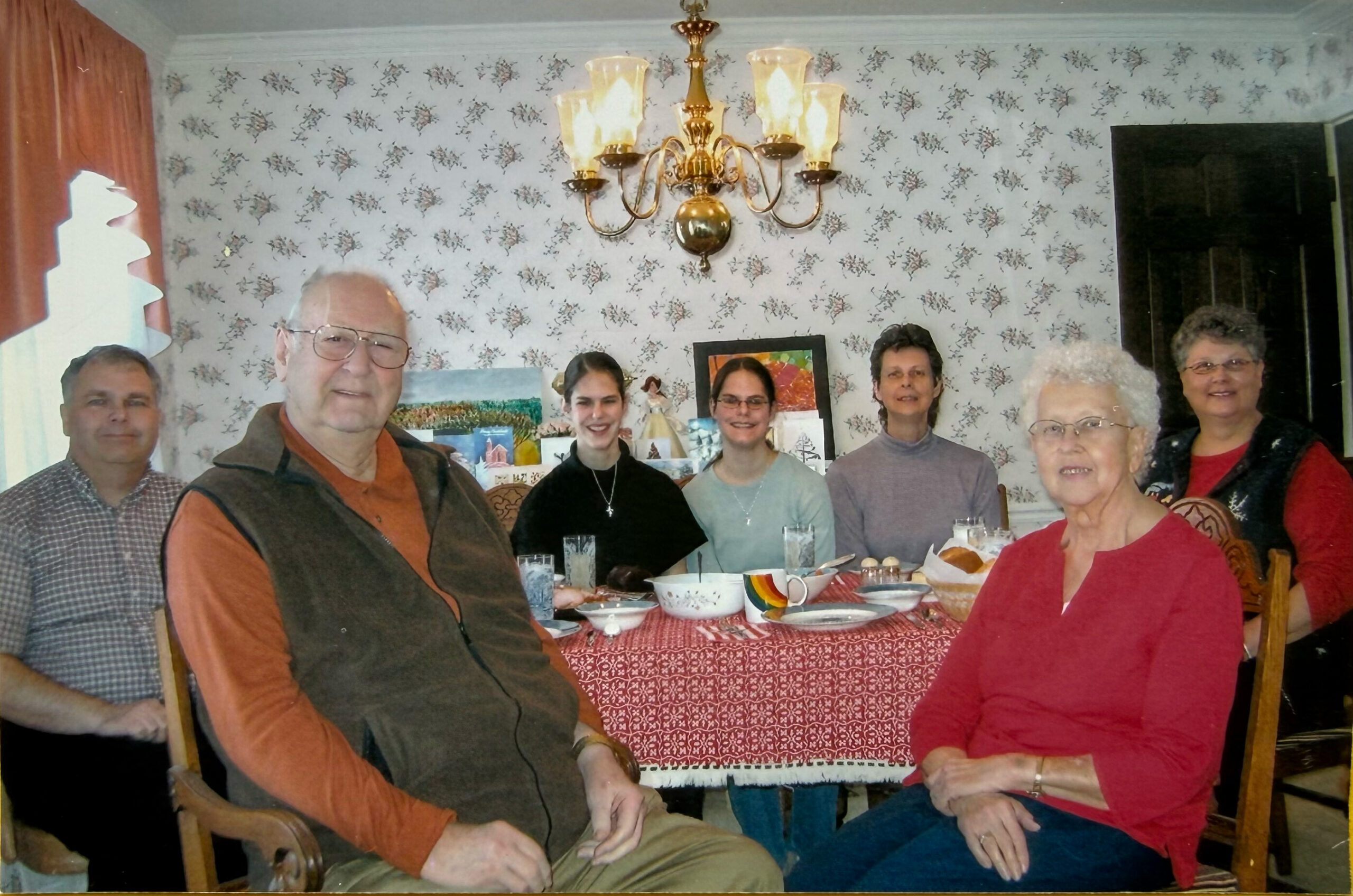 Group of seven adults sitting around a dining table with bowls and mugs, in a room with floral wallpaper and a brass chandelier above.