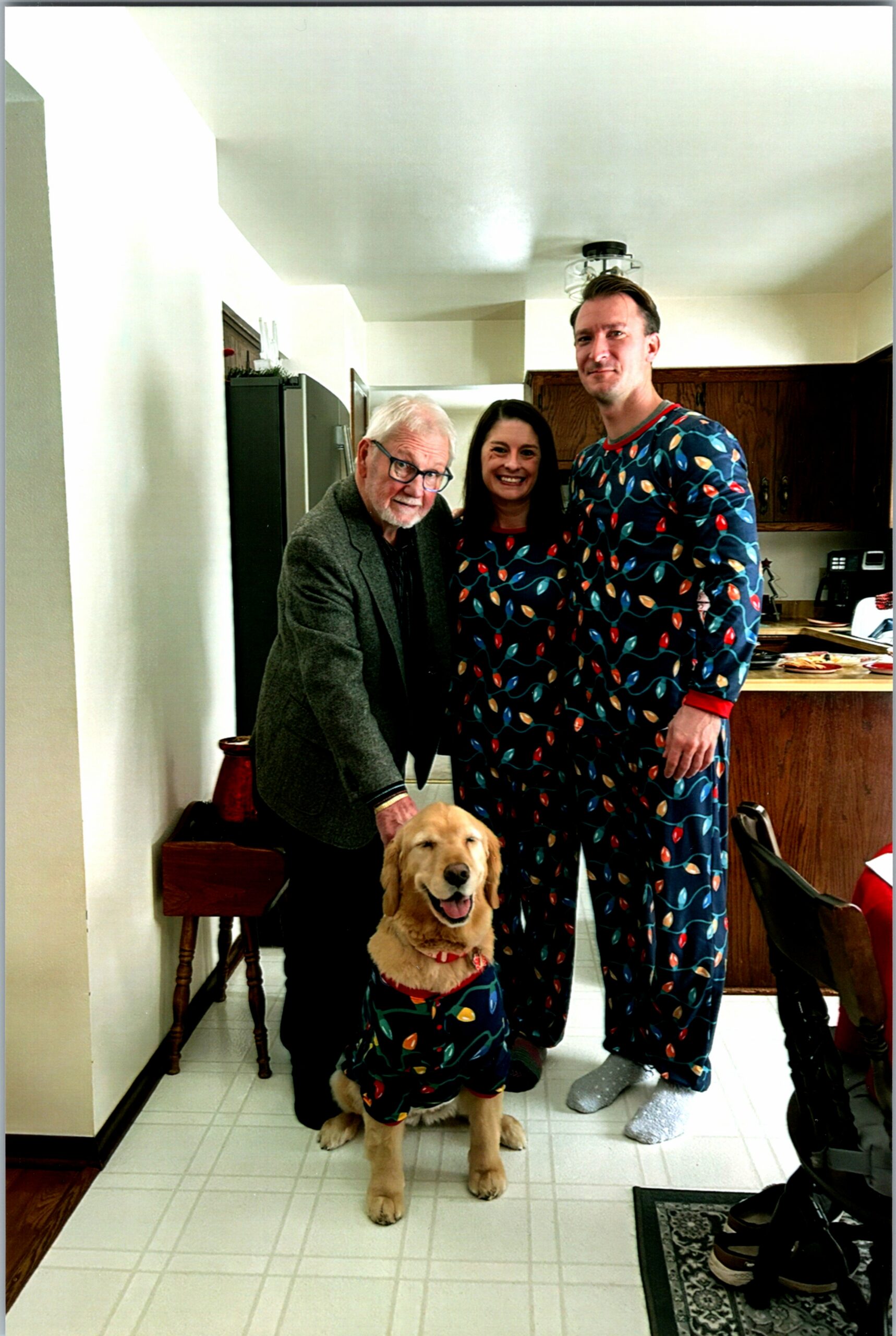 Family group in a kitchen posing for a photo with a golden retriever wearing matching holiday-pattern pajamas next to them.