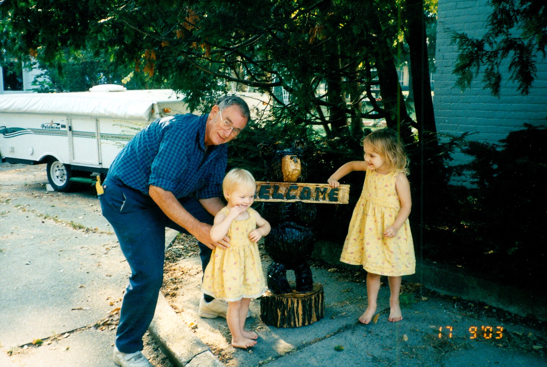 Older man in a blue plaid shirt leans toward two little girls in matching yellow dresses, posing beside a wooden 'WELCOME' sign on a stump outdoors with a trailer in the background.