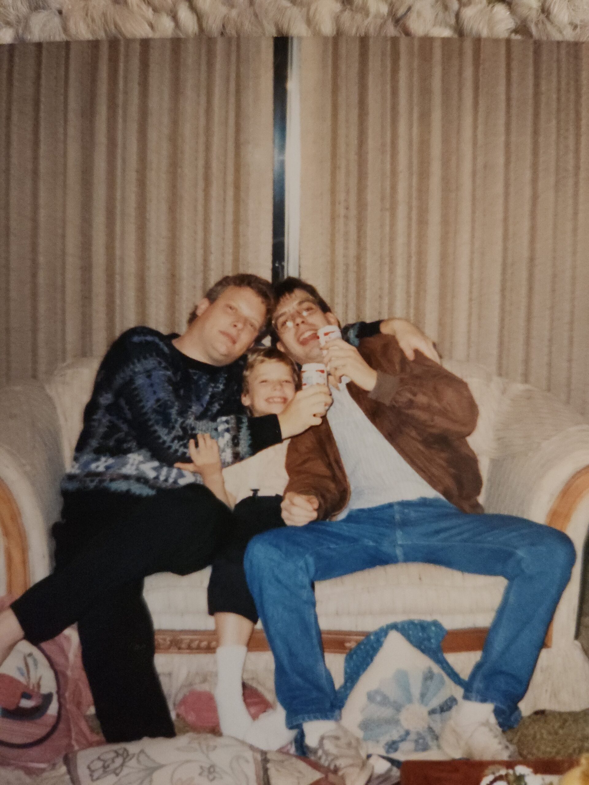 Three people—two adults and a child—sitting close on a beige couch, smiling and holding drinks in a cozy living room setting.