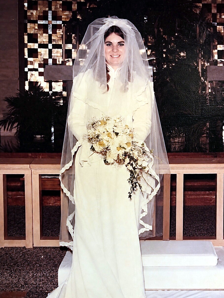 Bride in a white wedding gown with veil, holding a cream bouquet, indoors and smiling.