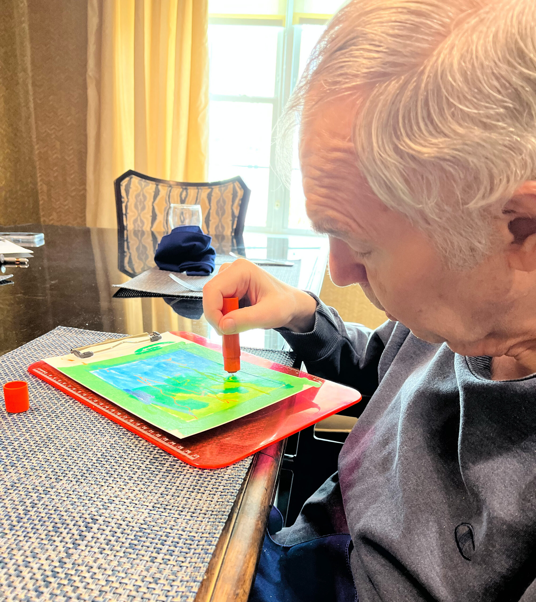 Close-up of a child’s hand using an orange marker to color a bright, abstract drawing at a dining table.