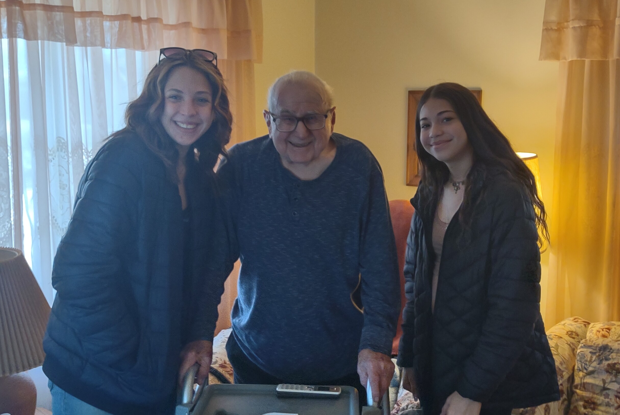 Three people posing together indoors: an older man between two young women, all smiling, with a printer and a phone on the table in front of them.