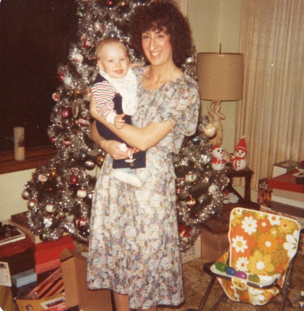 Woman in a floral dress holding a smiling baby in front of a decorated Christmas tree with gifts and festive decor in a living room.