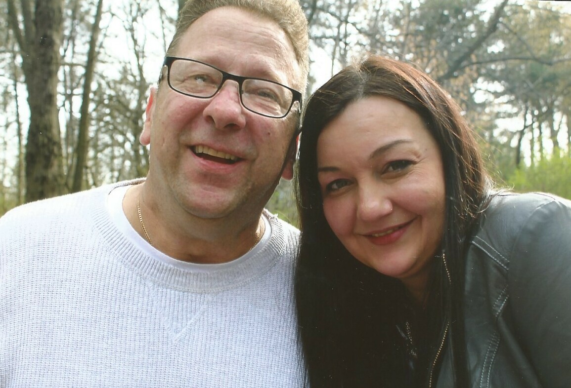 Smiling couple taking a selfie outdoors in a wooded area, posing close together for the photo.