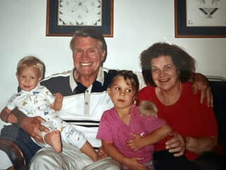 Smiling grandfather and grandmother sit with four young children on a couch, including a baby, in a cozy living room.