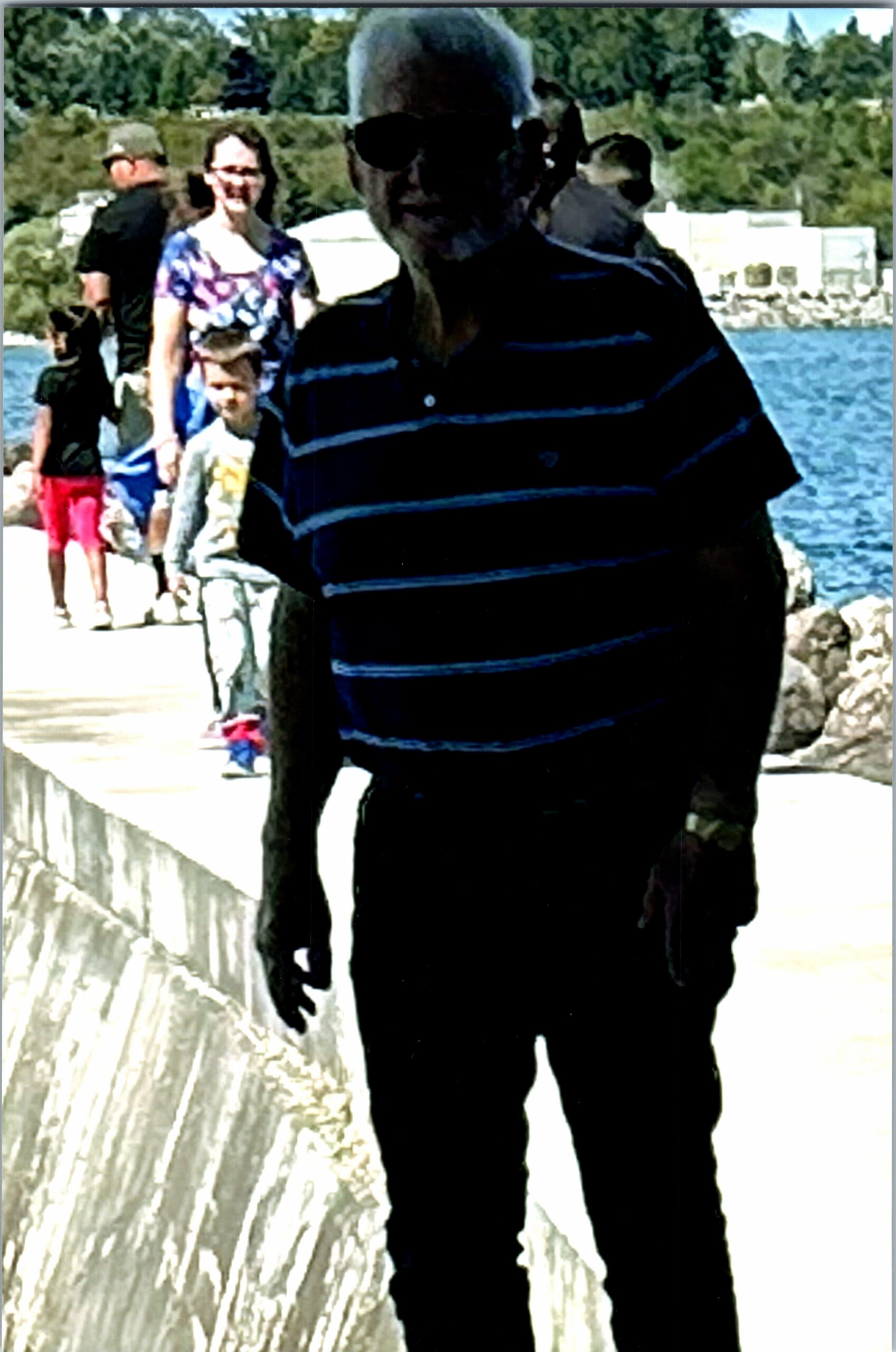 Man wearing dark, horizontally striped shirt and sunglasses stands near a stone railing by a waterfront as people walk along a path behind him.