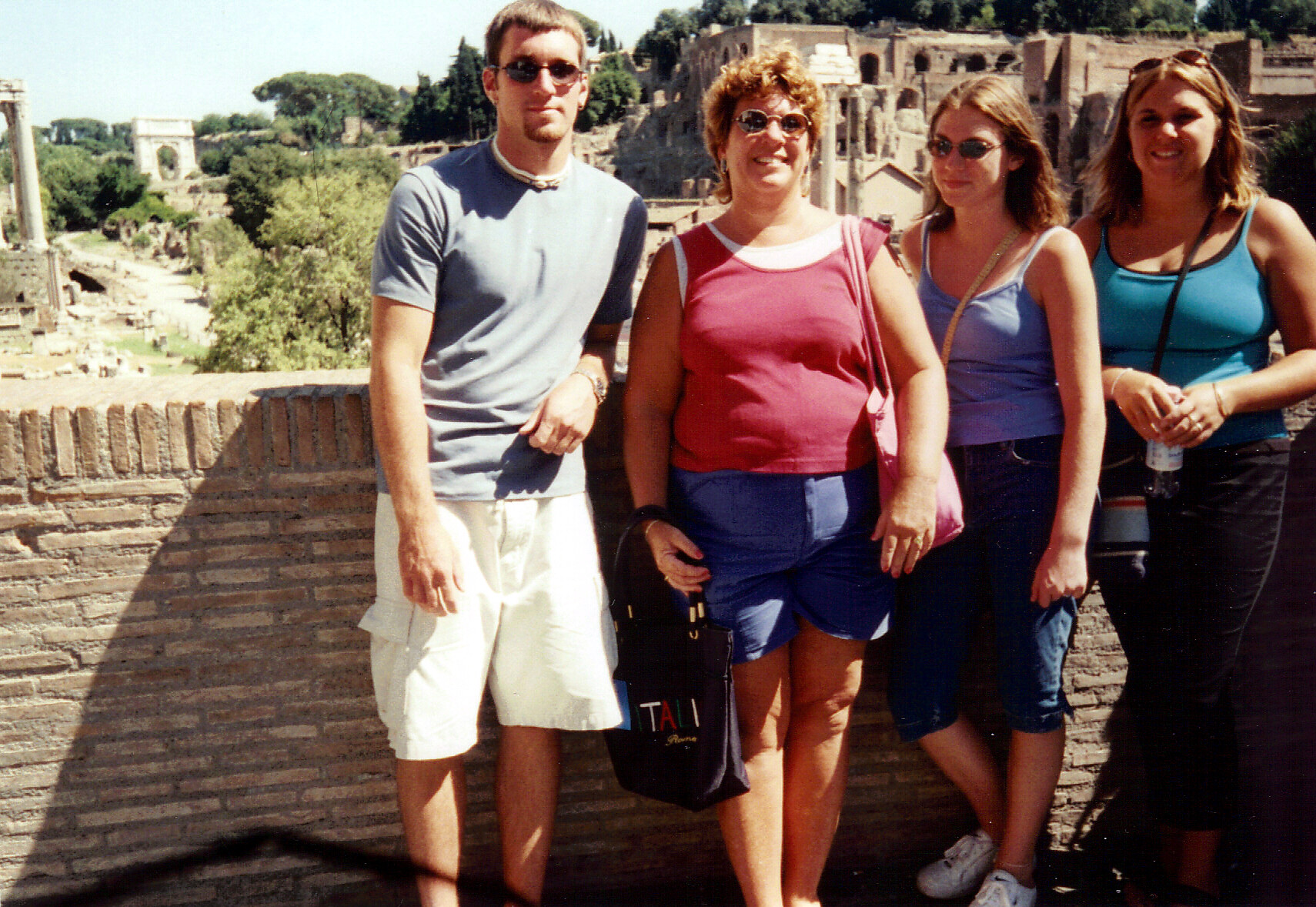 Group of four adults posing on a stone overlook with ancient ruins in the background on a sunny day.