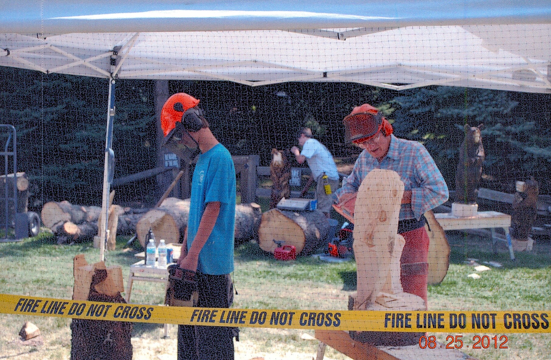 Two people under a white canopy and a table with supplies, yellow 'Fire Line Do Not Cross' tape in the foreground.