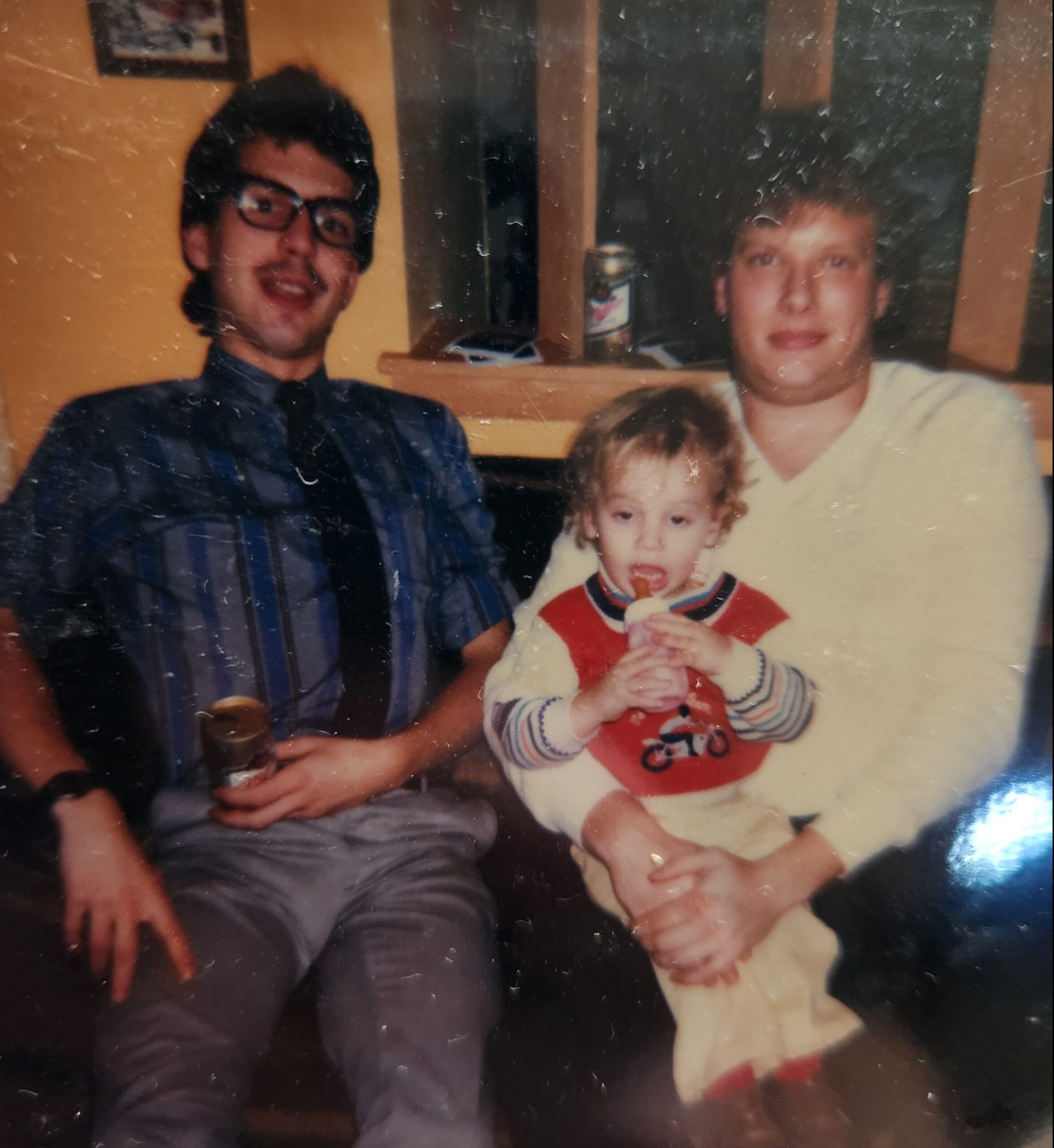 Three people posing indoors for a family photo: a man with glasses on the left, a man in a pale sweater on the right, and a toddler in front drinking from a bottle.