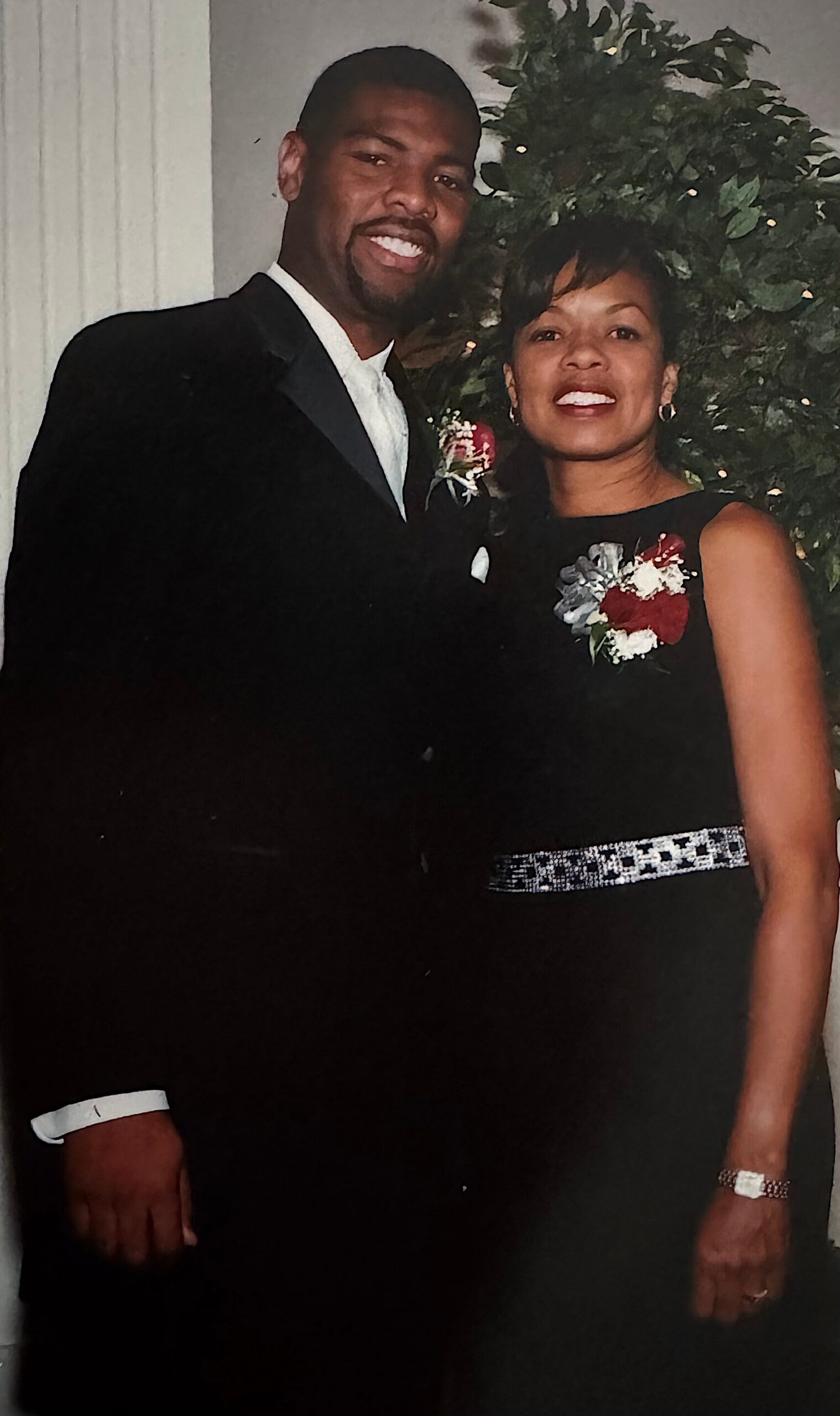 Smiling couple in formal attire posing indoors with a greenery backdrop, the man in a tuxedo and the woman in a black dress with corsages.