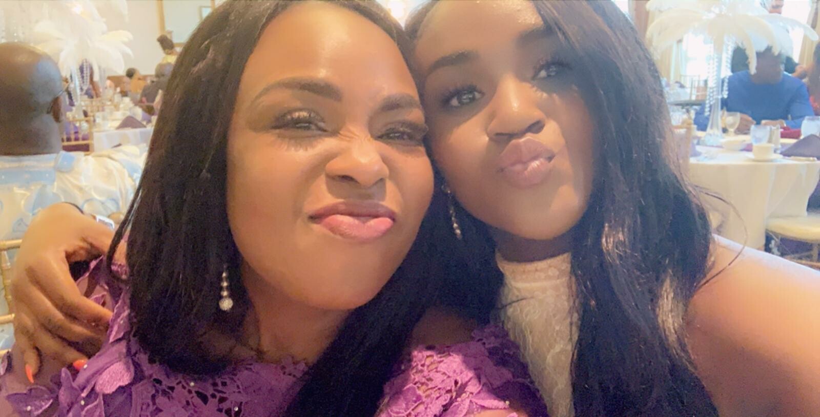 Two women pose for a selfie at a formal banquet, one in purple lace and the other in white, smiling with heads close together; white feather centerpieces and tables in the background.
