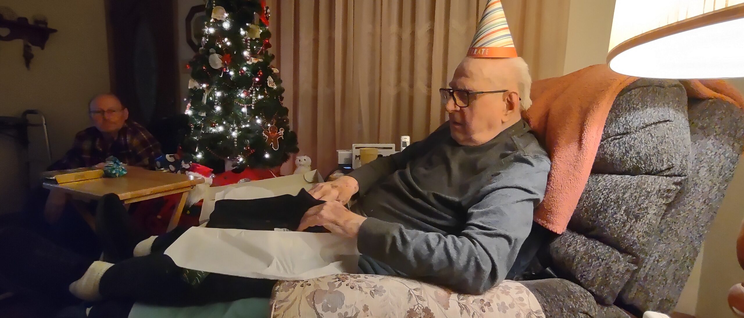 Elderly man in a gray shirt and glasses sits in a recliner with a party hat, holding hands with someone off-camera, in a living room with a decorated Christmas tree and presents.