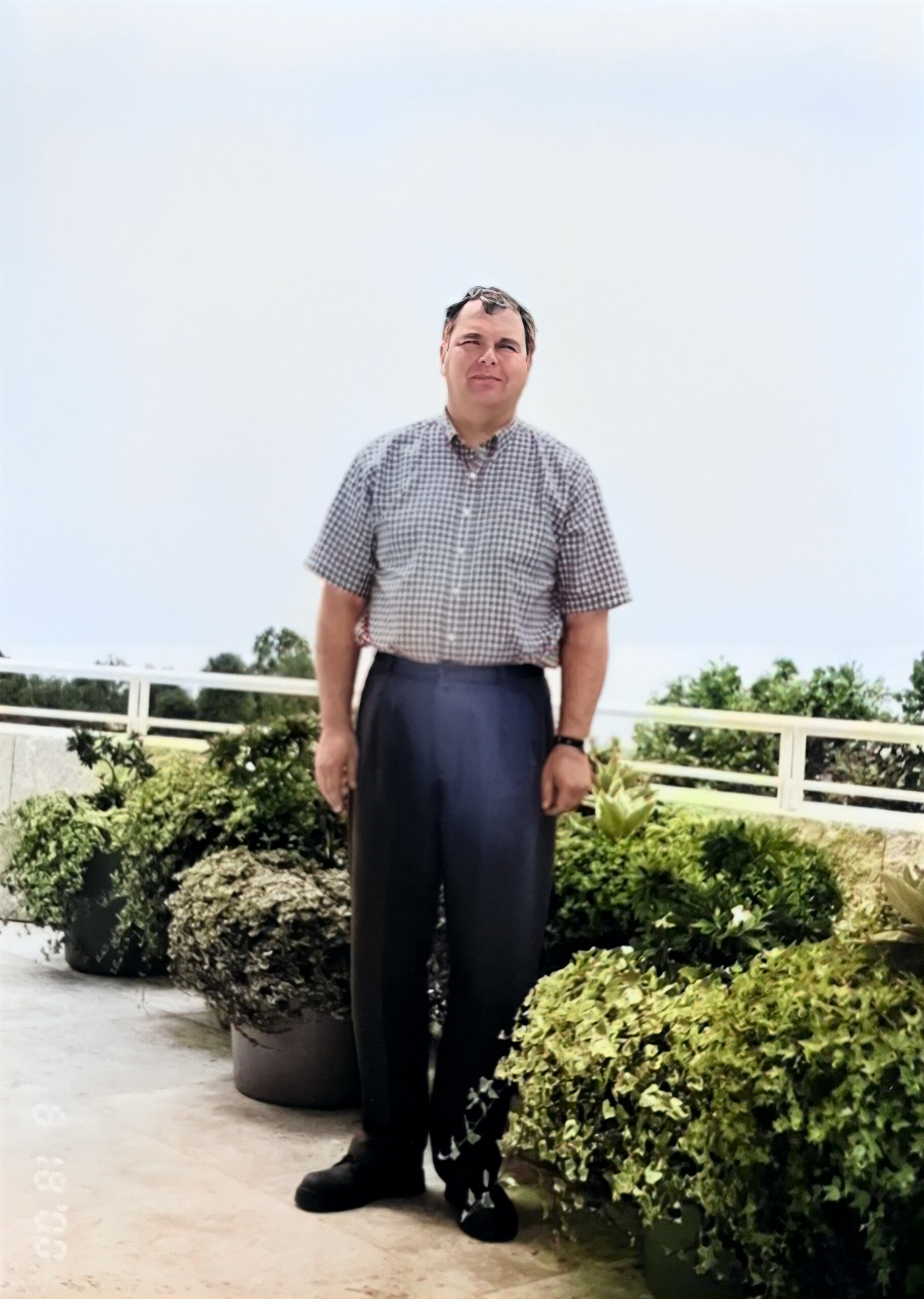 Man standing outdoors on a balcony/terrace with potted plants, wearing a short-sleeve checkered shirt and dark trousers.