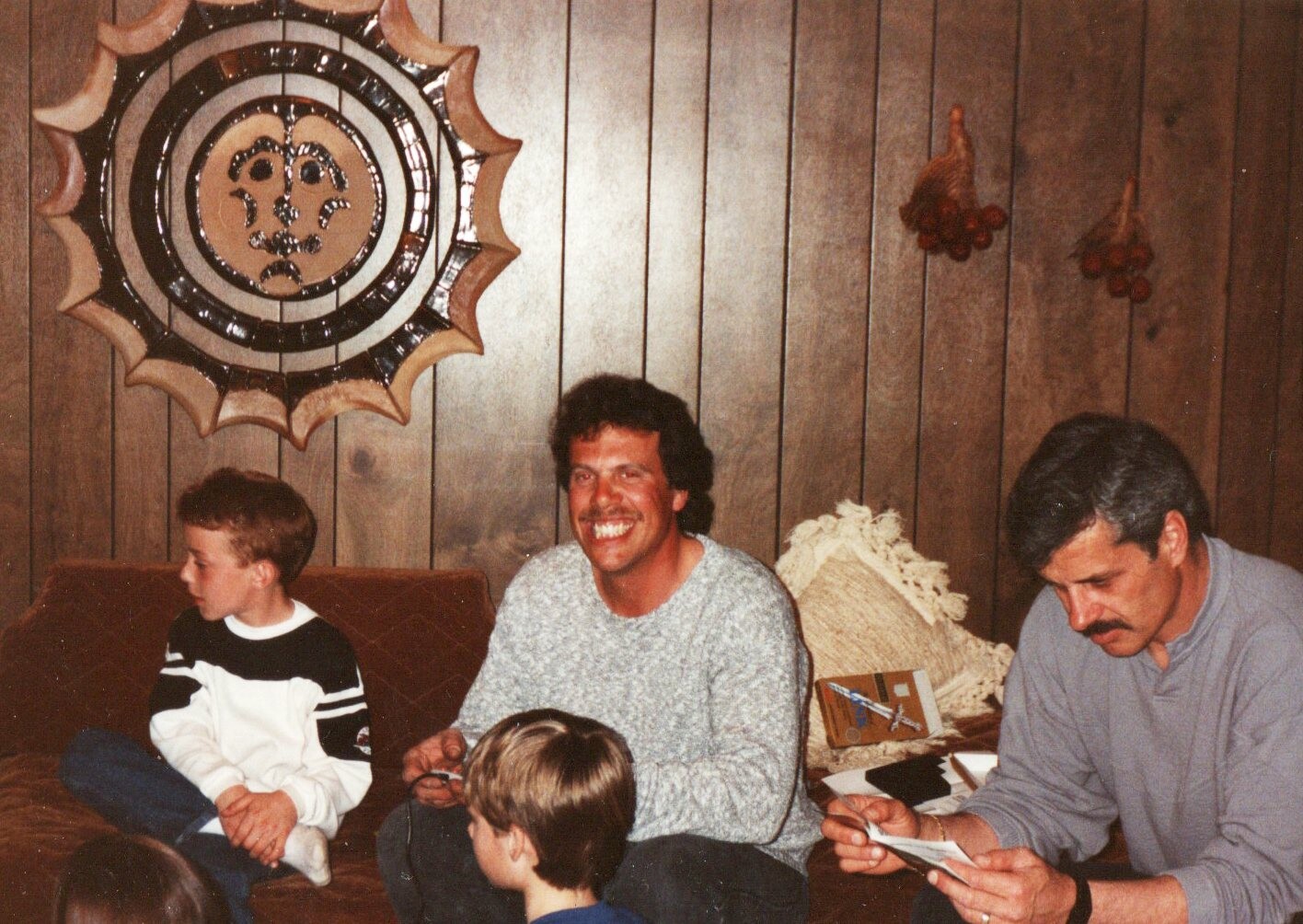 Family sits on a brown couch in a wood-paneled living room; a smiling man sits center with two boys nearby, another man reads papers on the right.