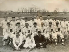 Black-and-white photo of a vintage baseball team posing on a dusty field, players in uniforms with an S on the chest and a man in a dark suit kneeling in front center creating a two-row group portrait.