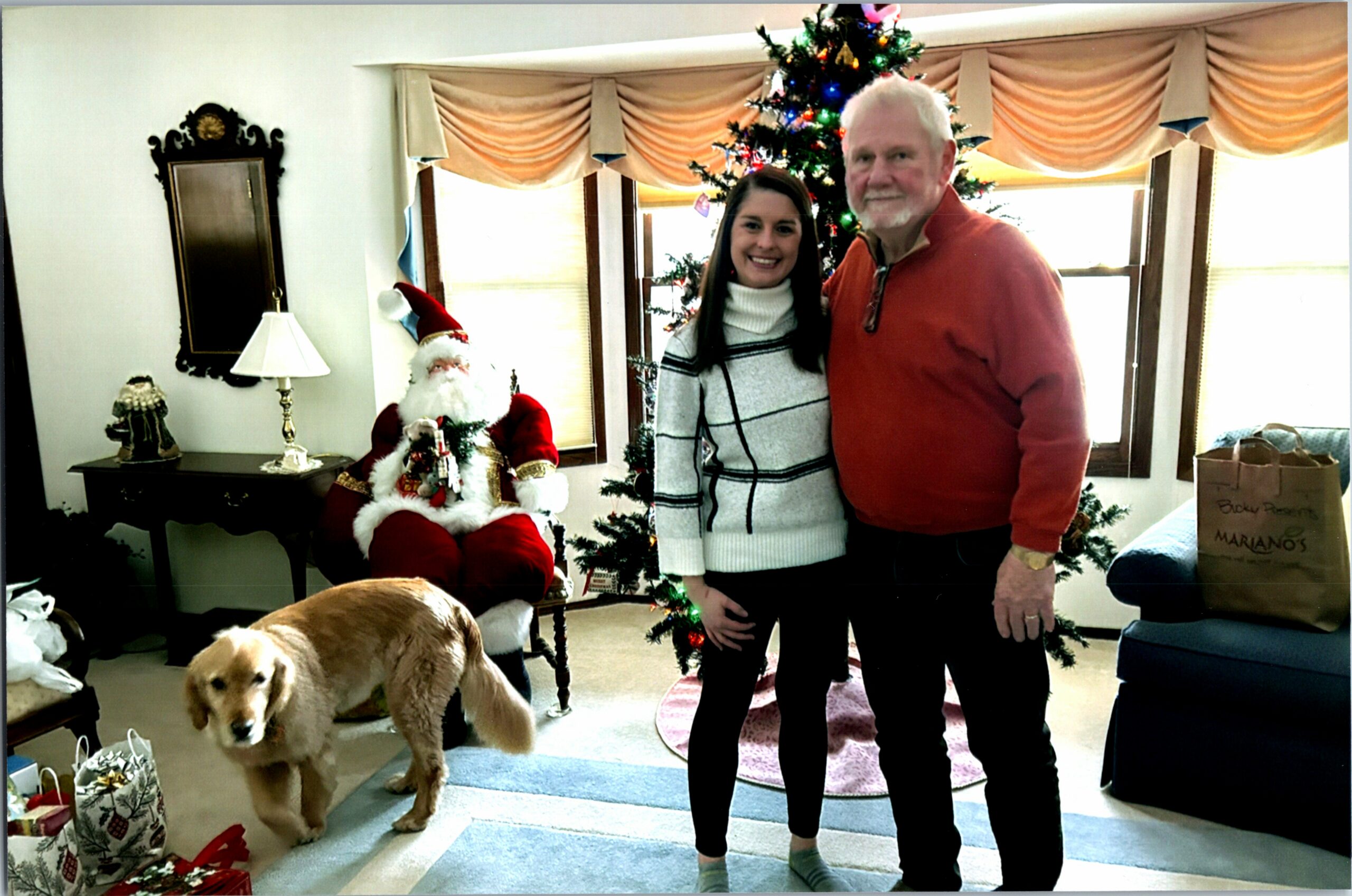 Two adults posing by a Christmas tree with Santa Claus, a golden retriever exploring gifts nearby in a cozy living room.