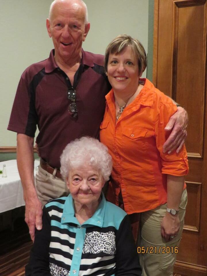 Elderly woman seated in a teal and black striped cardigan, flanked by a man in a maroon polo and a woman in an orange shirt, all smiling indoors.