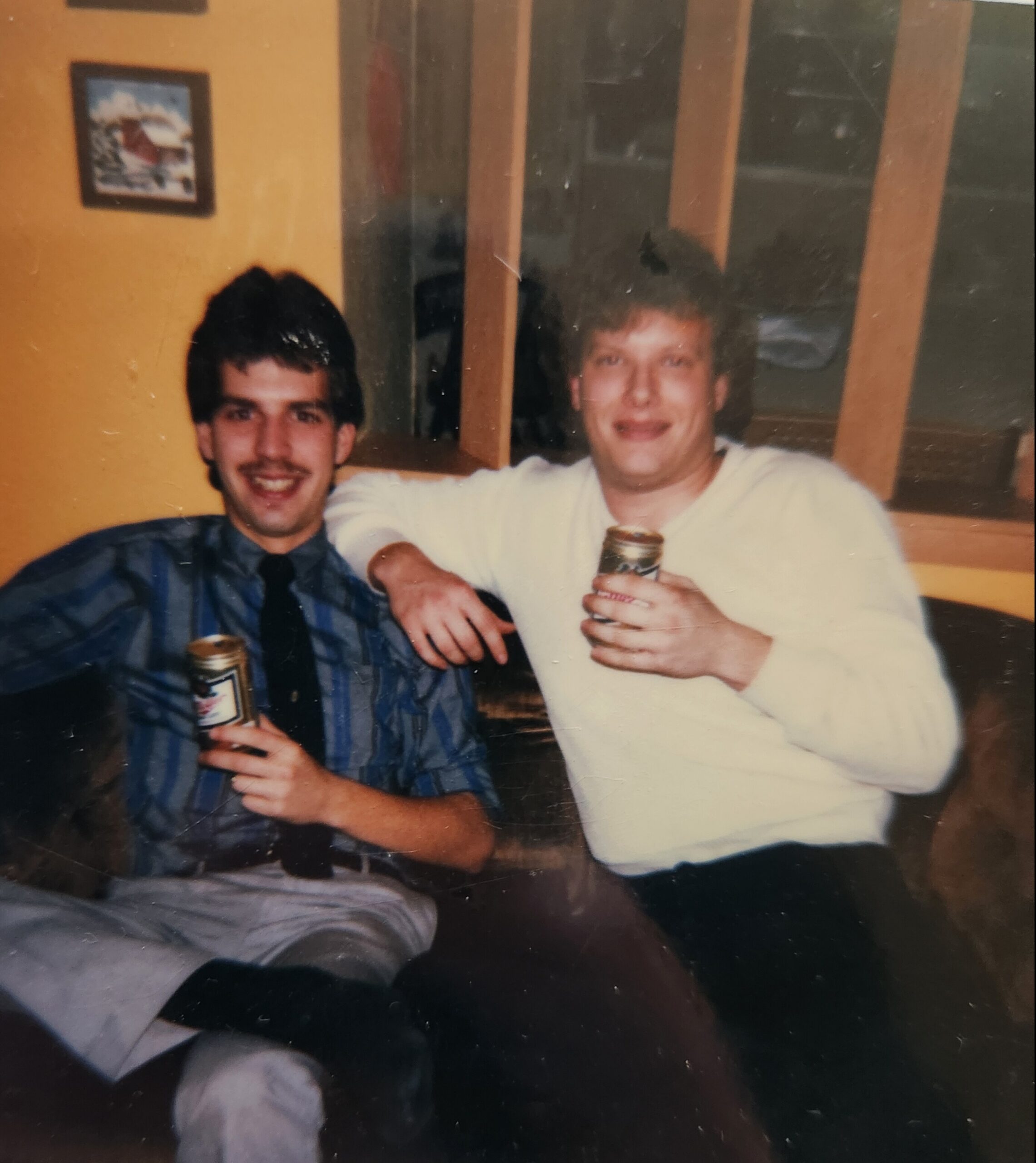 Two men pose for a casual photo indoors, each holding a beer bottle; one in a blue striped shirt, the other in a light sweater, against an orange wall.