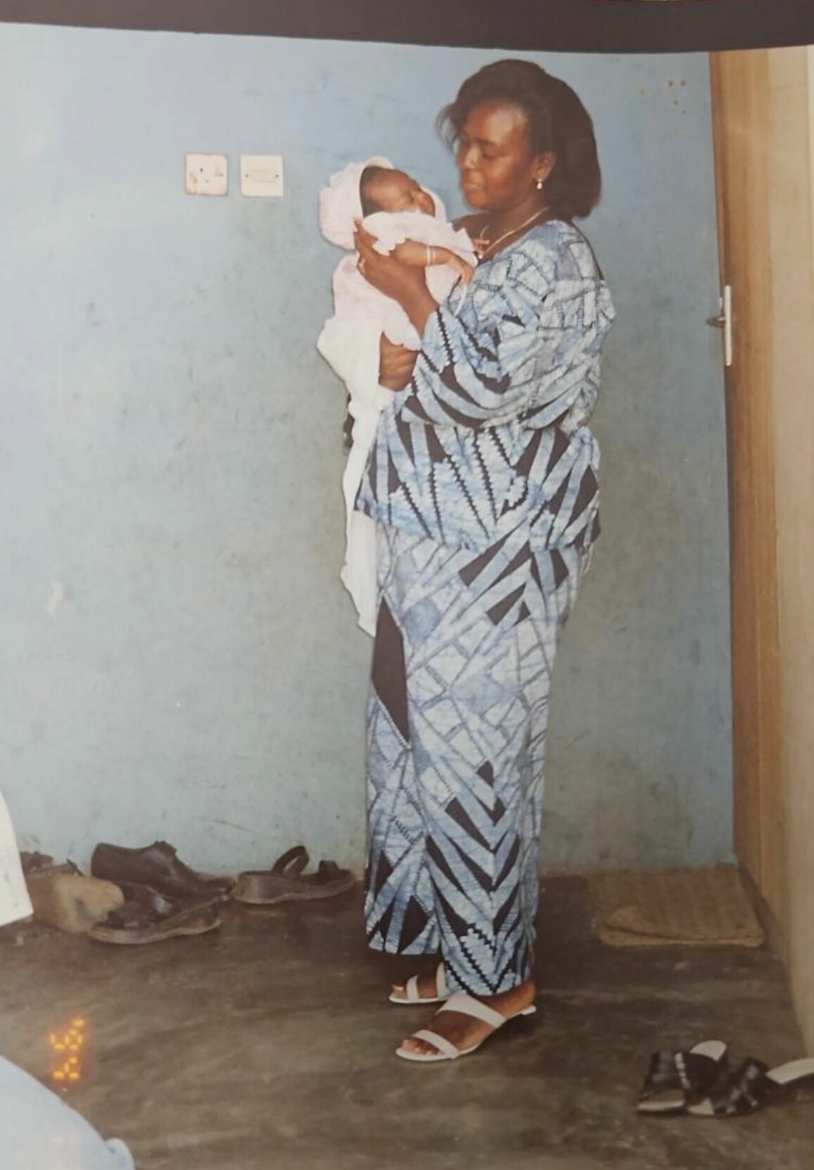 Woman in a blue patterned dress gently holding a newborn wrapped in white, indoors by a blue wall.