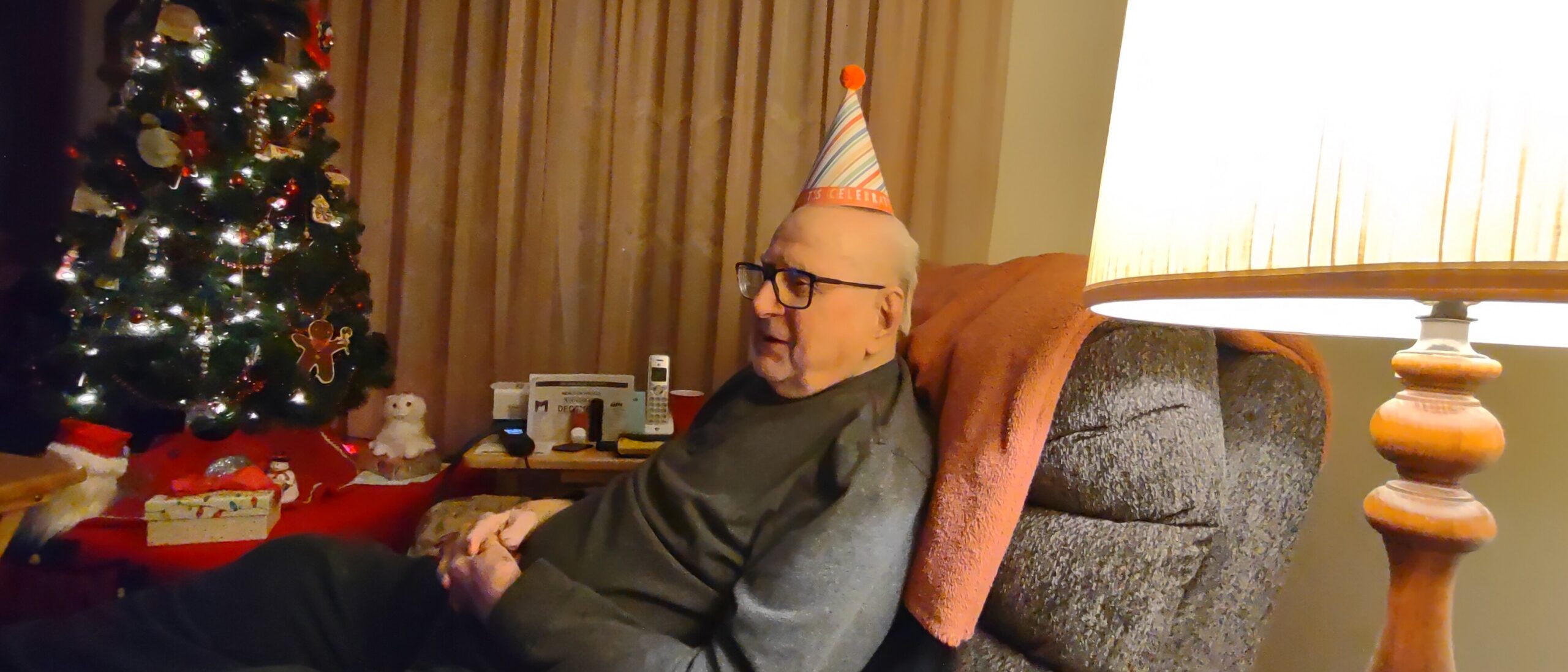 Older man wearing a striped party hat sits on a sofa beside a lit Christmas tree and wrapped gifts in a living room.