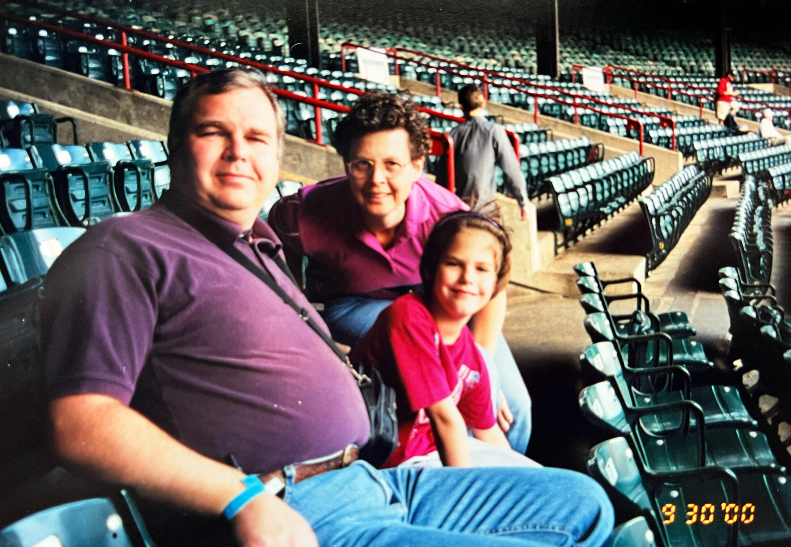 Three family members pose for a photo in an empty stadium with teal seats and red railing, the man on the left, a woman and a girl in the foreground smiling at the camera.