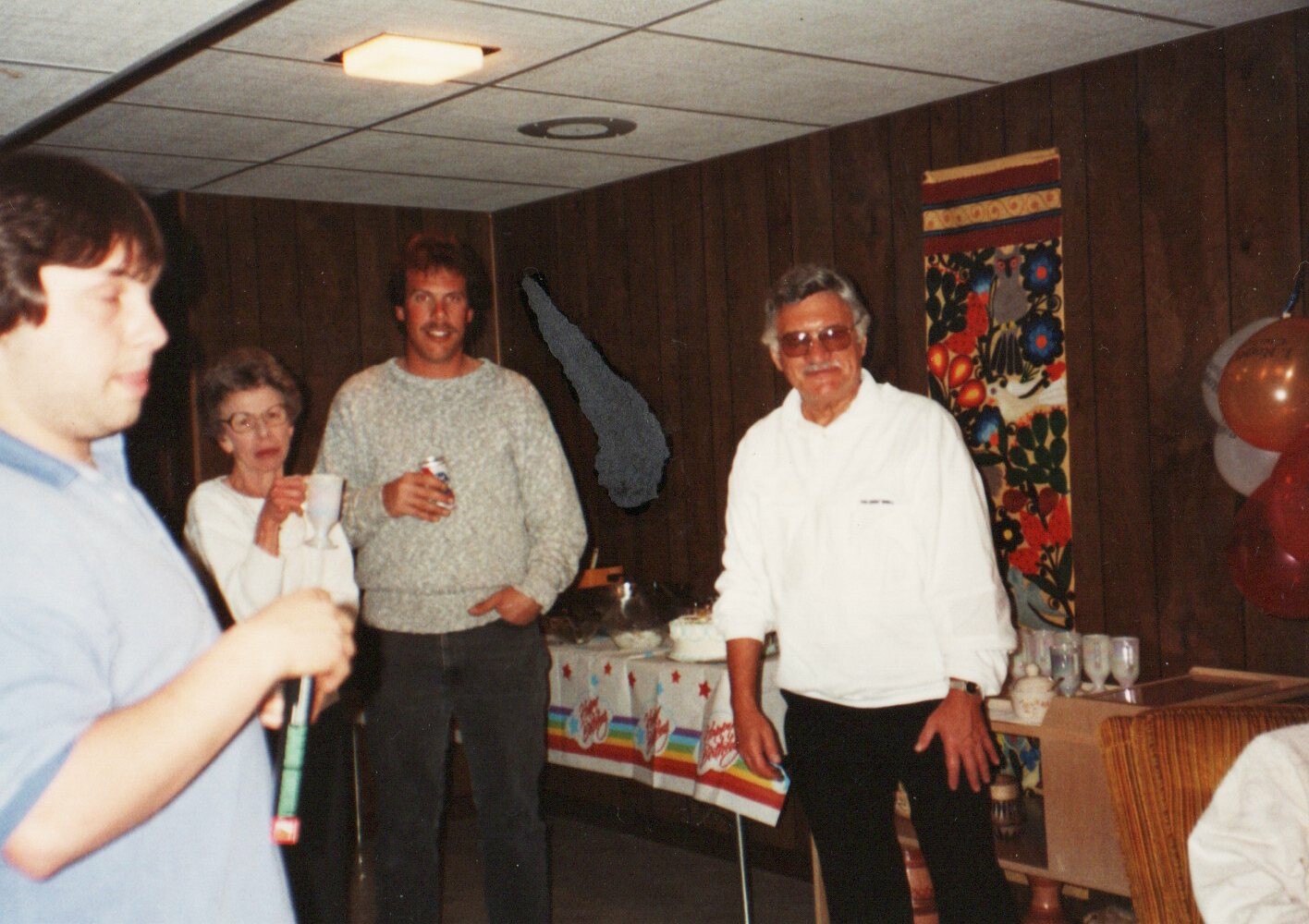 Group of people at a casual indoor party in a wood-paneled room, with drinks in hand near a buffet table and balloons in the corner.
