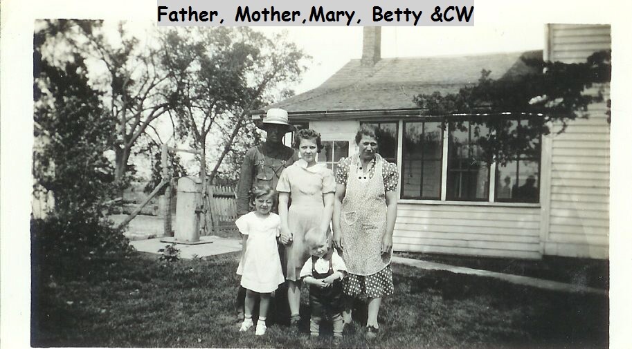 Family of five posing on a lawn in front of a house: father wearing a hat, mother in an apron, two daughters, and a toddler.