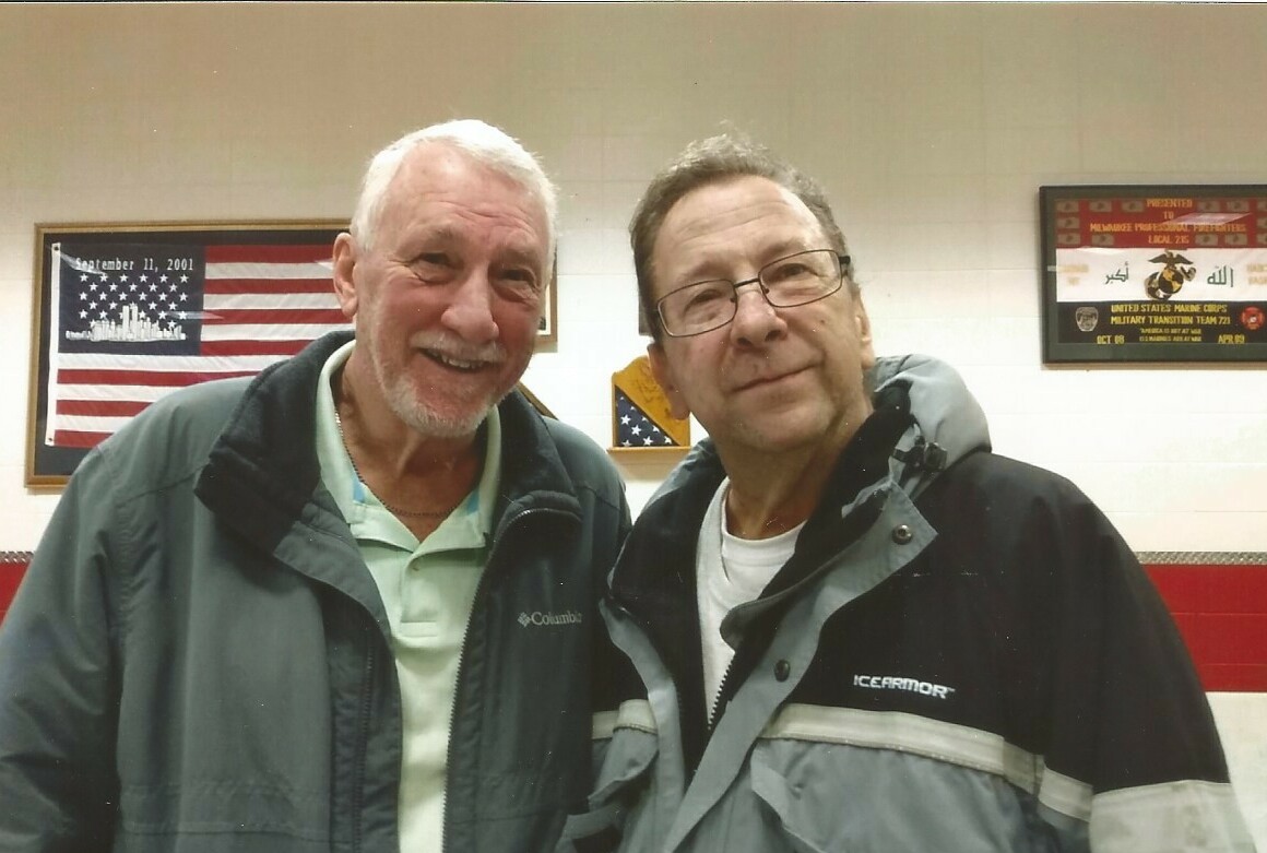 Two smiling men in jackets pose together indoors; an American flag hangs on the wall behind them.