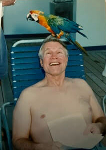 Smiling shirtless man lounging on a blue pool chair with a colorful parrot perched on his head outdoors.
