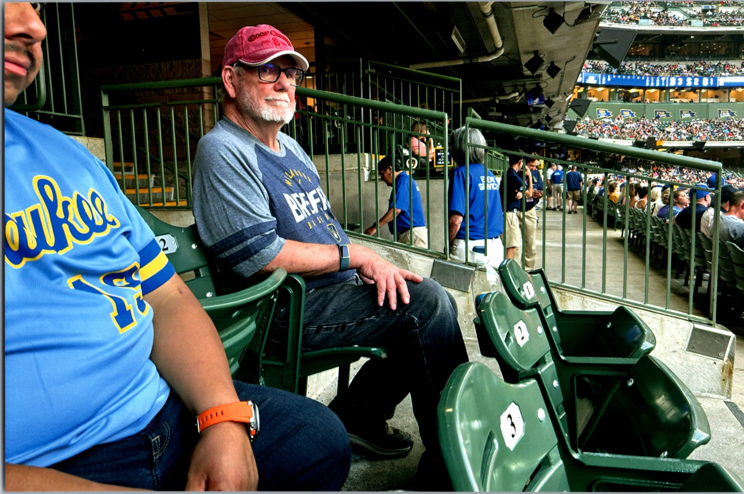 Man with a gray beard wearing a red cap and glasses sits in stadium seats, looking toward the field as a crowd fills the stands behind him.