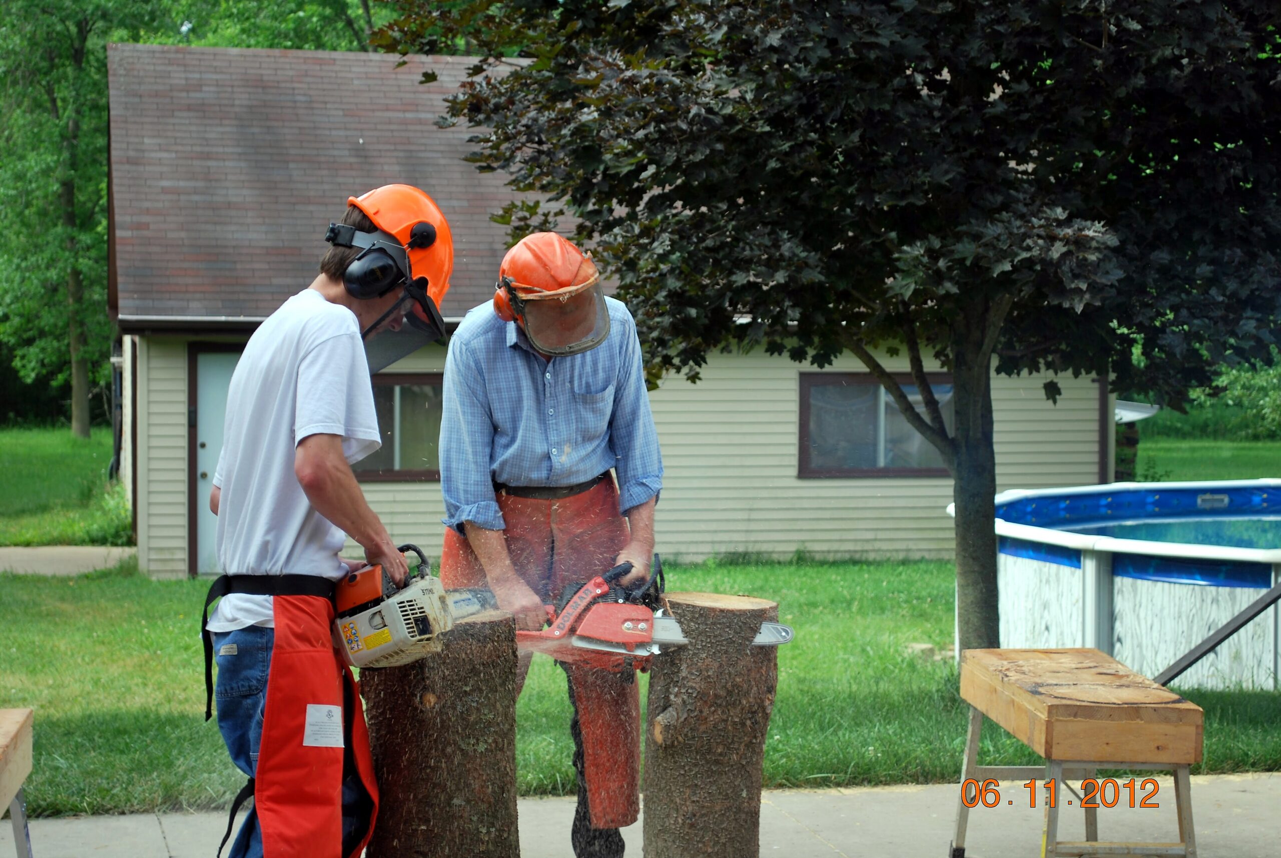 Two men in safety gear using chainsaws to cut a log outdoors in a yard under a tree.