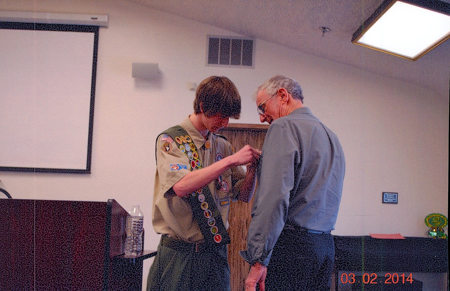 Scout in uniform pins a badge onto an older man in a blue shirt in a classroom setting with a water bottle on the counter nearby.