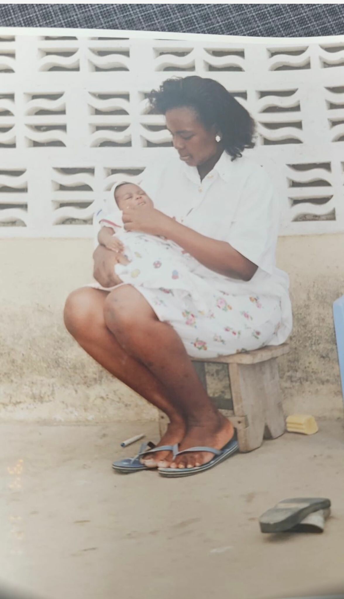 Woman sitting on a wooden stool, cradling a newborn wrapped in a floral blanket outdoors.