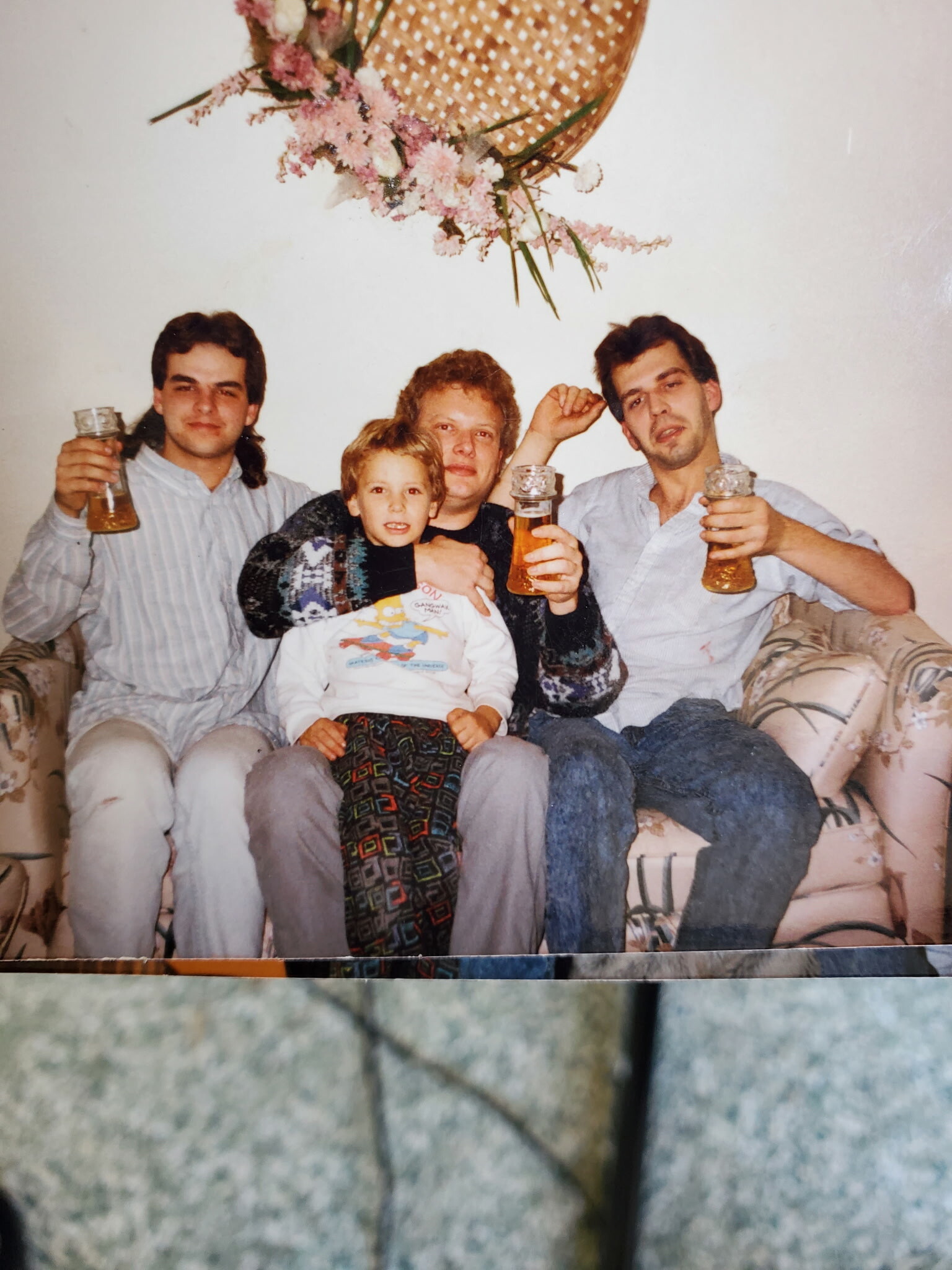 Four people sit on a patterned couch, raising beer glasses in a casual toast, with a child on the woman's lap, all smiling at the camera.