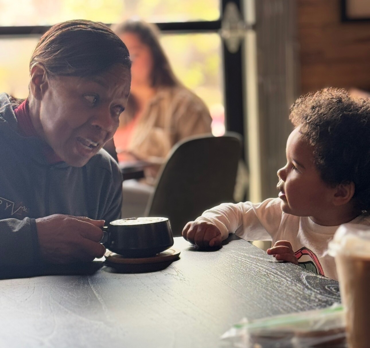 Adult and young child share a friendly conversation at a cafe table, with a mug near them and warm light in the background.