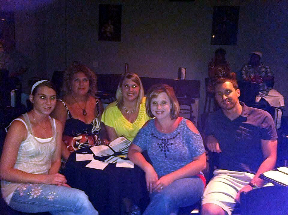 Five adults pose for a photo at a dimly lit table during a casual gathering; papers and napkins are on the table in front of them.