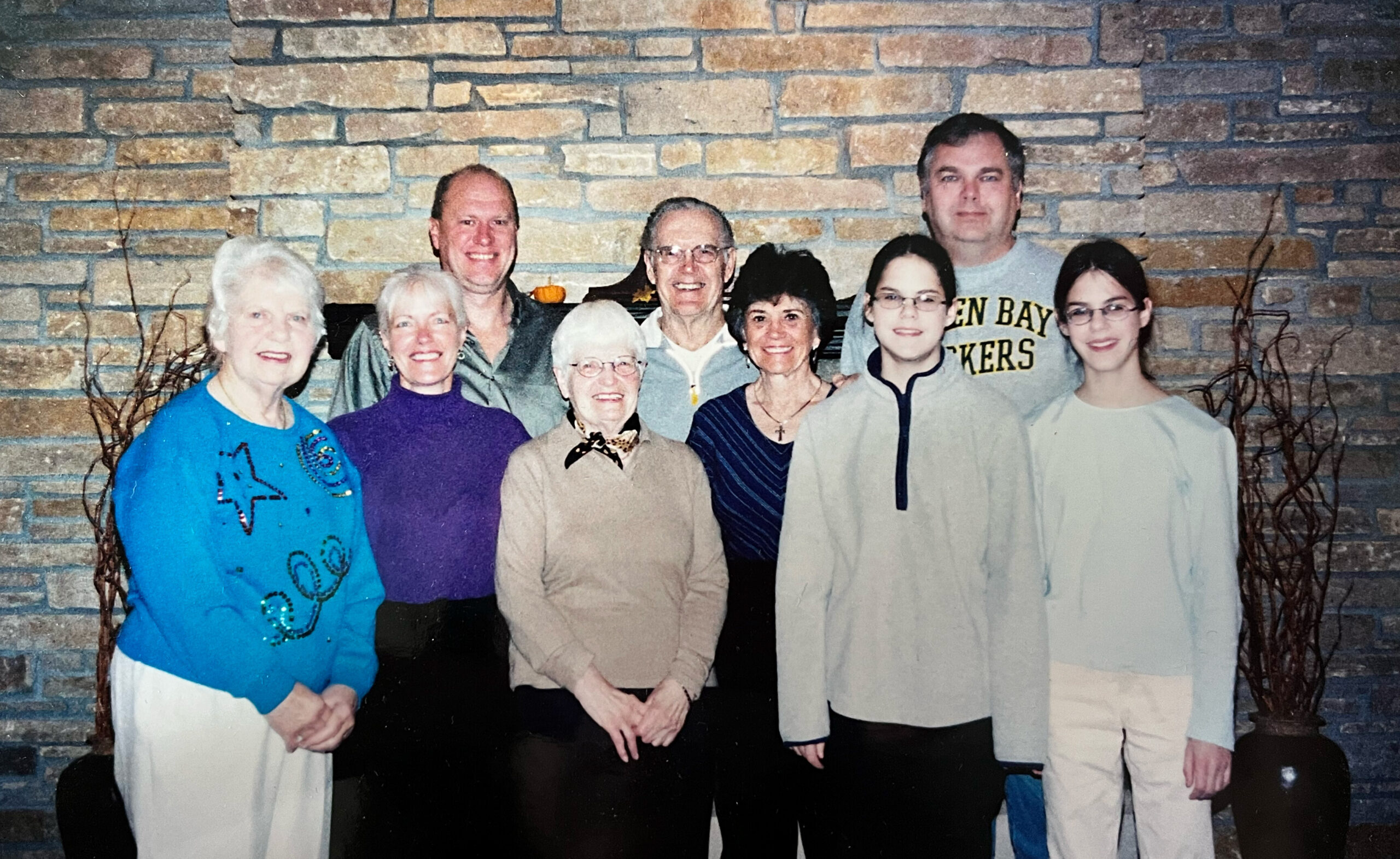 Nine people posing for a group photo indoors in front of a brick fireplace, all smiling at the camera.