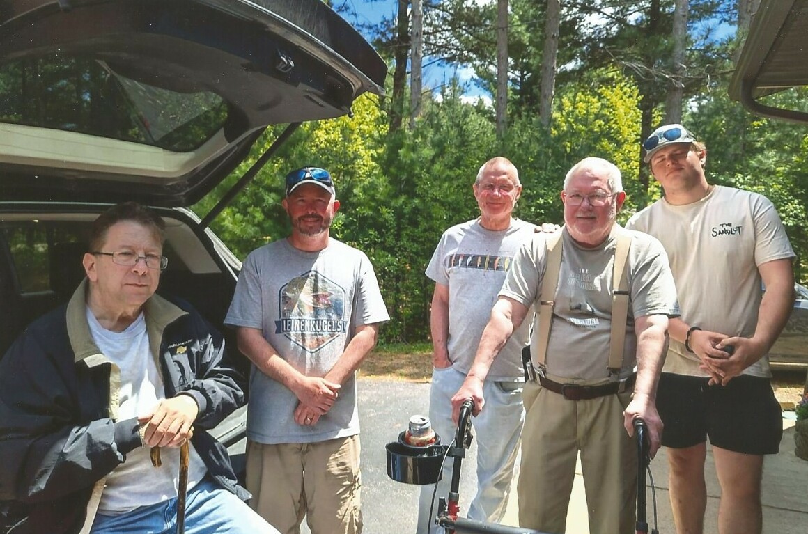 Five men stand together on a sunny driveway near an SUV with the trunk open, trees in the background.