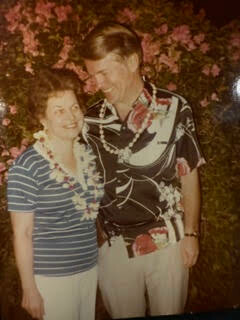 Smiling couple standing close together at night, both wearing flower leis; woman in striped shirt and white shorts, man in tropical-pattern shirt, pink flowers in background.