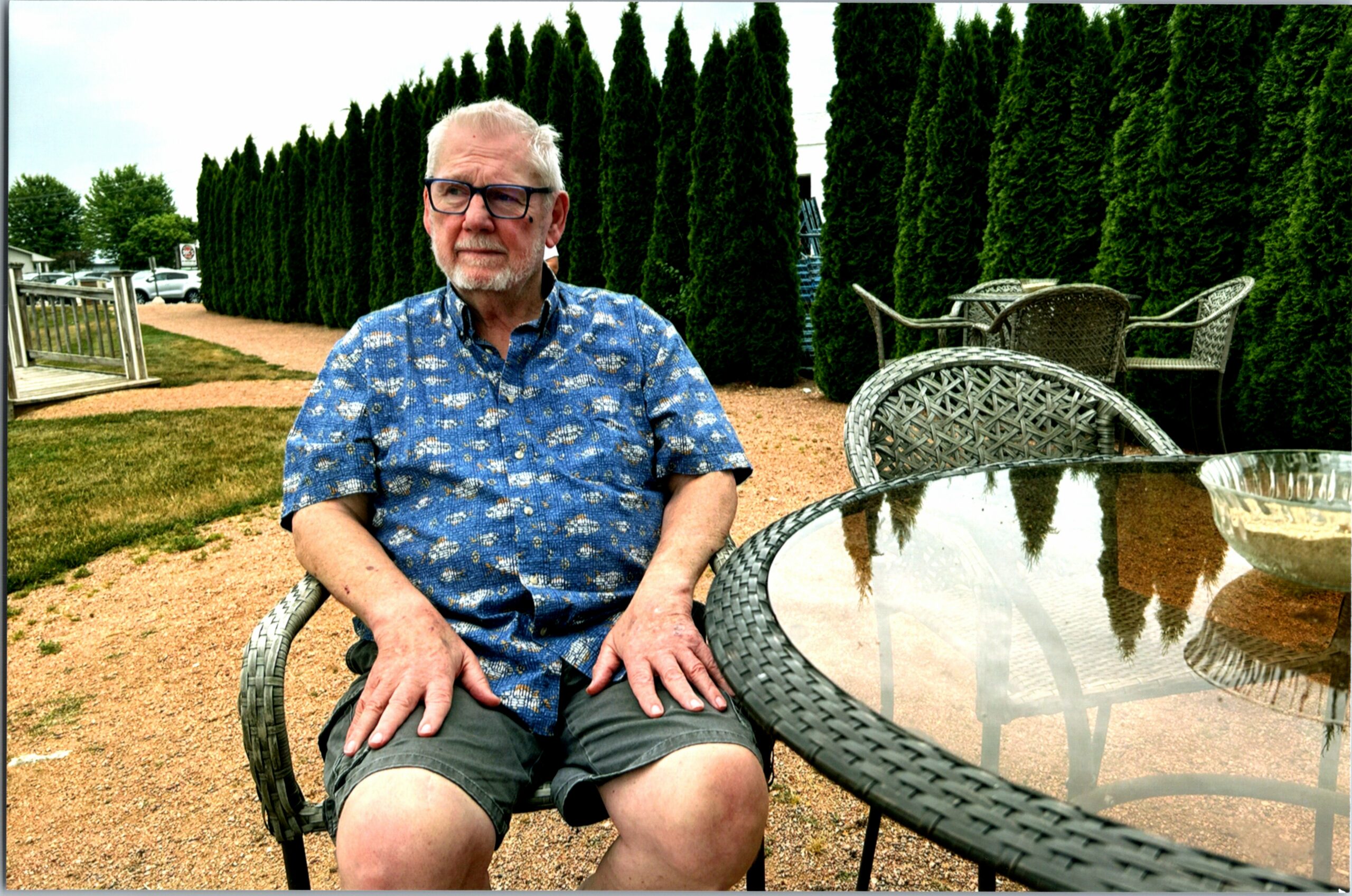 Older man with gray hair and glasses sits at an outdoor wicker table, with tall evergreen hedges behind him.
