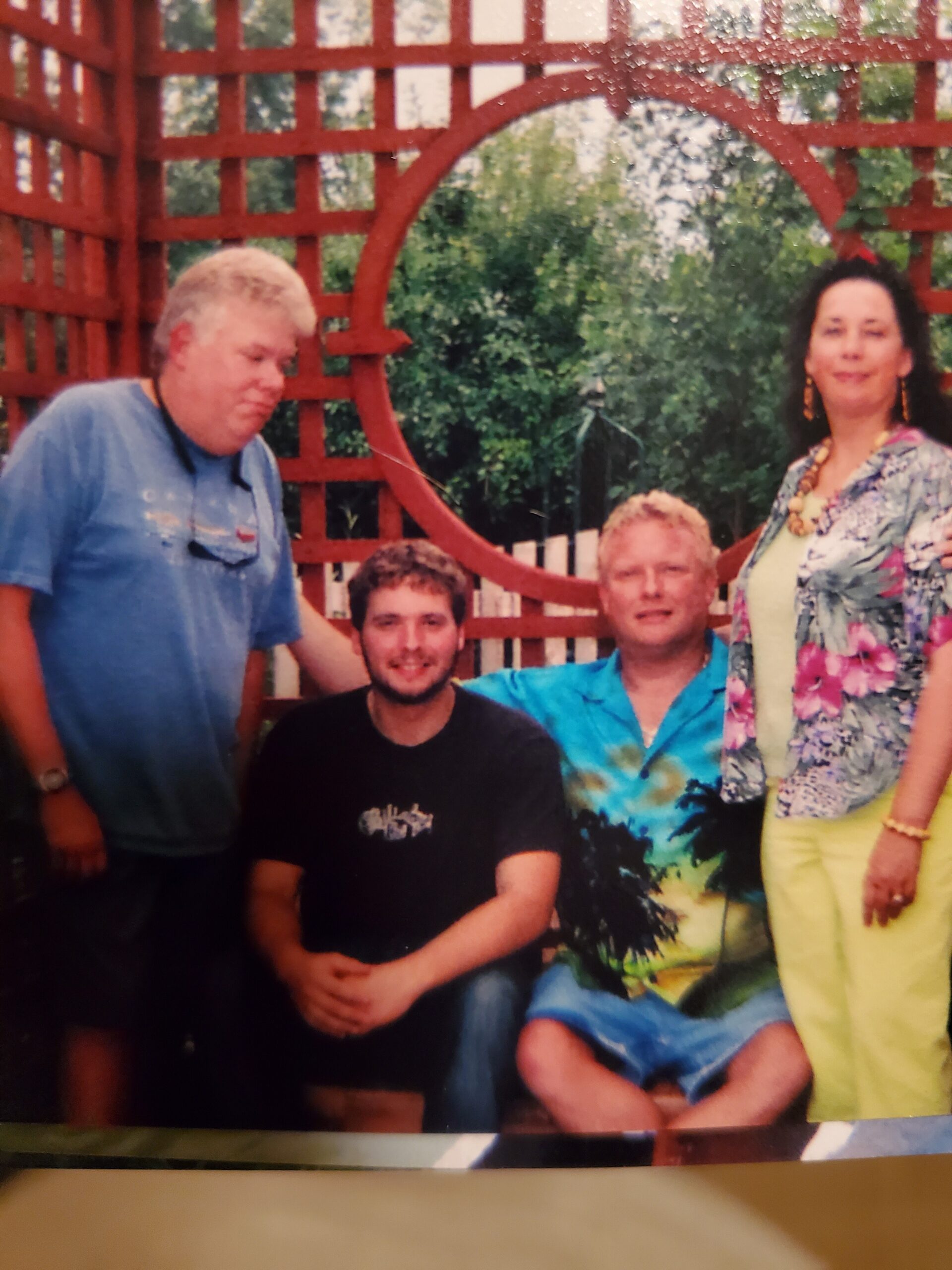 Four adults posing outdoors in front of a red wooden lattice arch, smiling at the camera with greenery in the background.