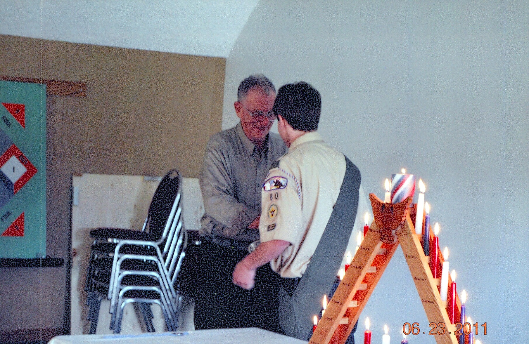 Two men shake hands near a tall wooden candle arch, one in a tan scout-like uniform and the other in a gray shirt, inside a room with stacked chairs in the background.