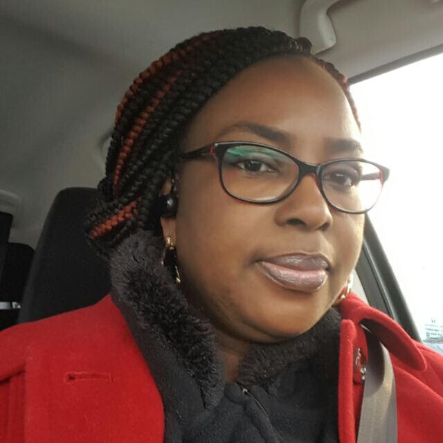 Close-up of a Black woman in a car, wearing red-framed glasses, red-tipped braids, and a red coat with a visible seatbelt.