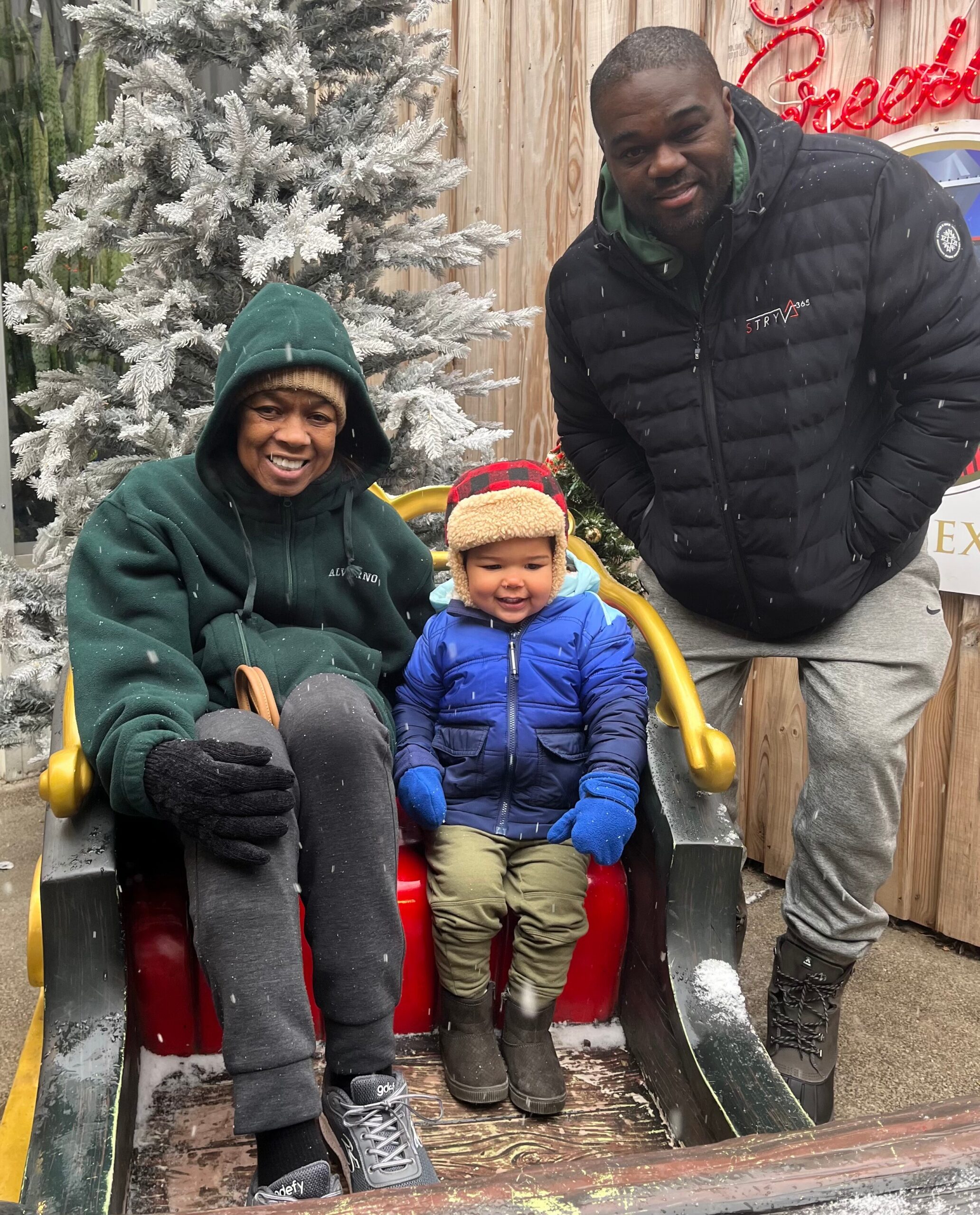 Woman in a green hoodie and a young child in a blue jacket sit together on a red sled, with a man in a black jacket standing nearby in a snowy outdoor scene with frosted trees and a wooden backdrop.