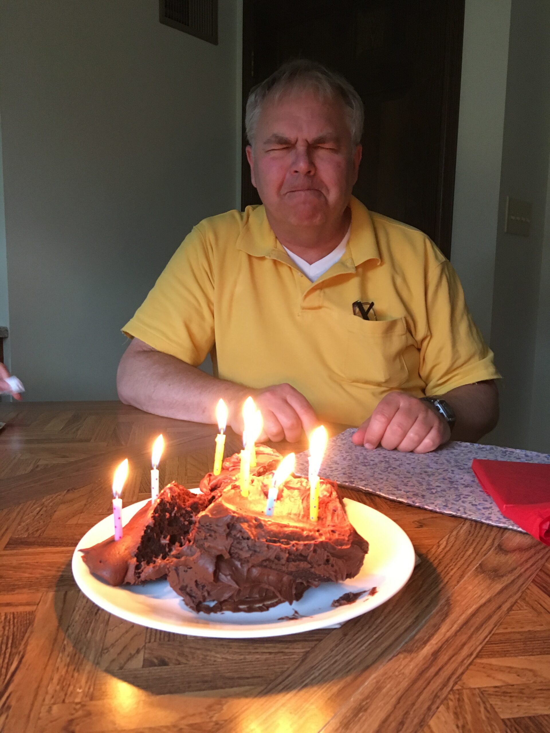 Older man in a yellow polo sits at a table in front of a chocolate birthday cake with lit candles.