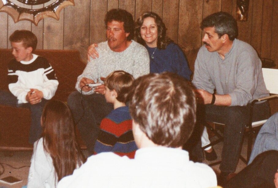 Family gathering in a wood-paneled living room; two adults sit on a couch with a child between them while kids gather around.