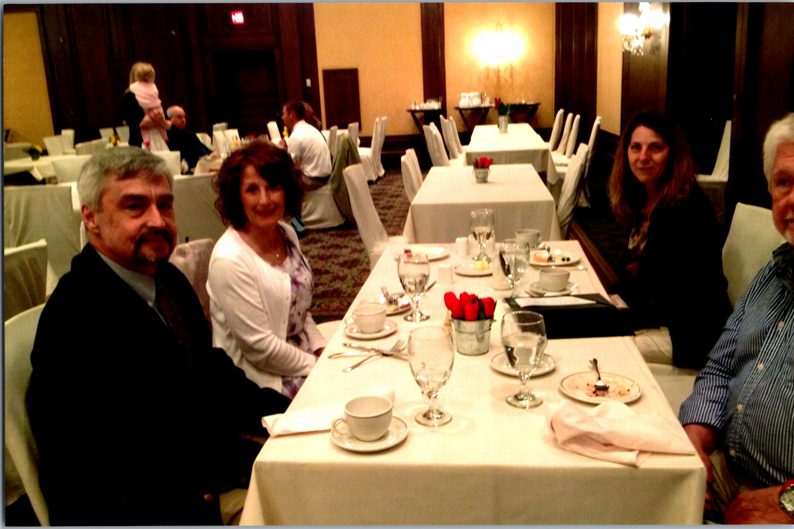 Group of adults dining together at a long, white-tablecloth restaurant table with dishes and wine glasses in a formal setting.