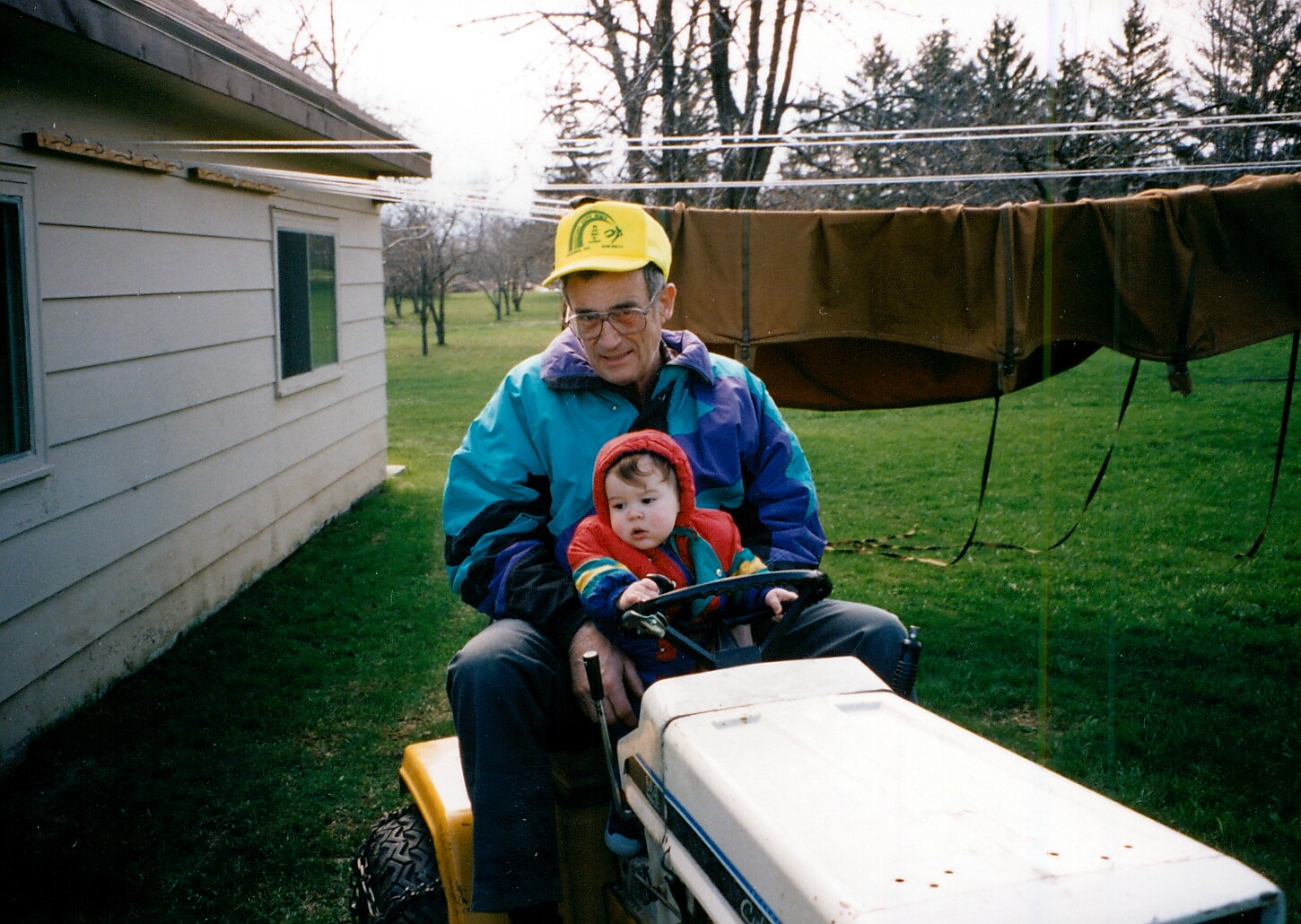 Older man in a blue and turquoise jacket with a yellow cap sits on a white riding lawn mower, guiding a toddler in a red hooded jacket in a backyard garden.