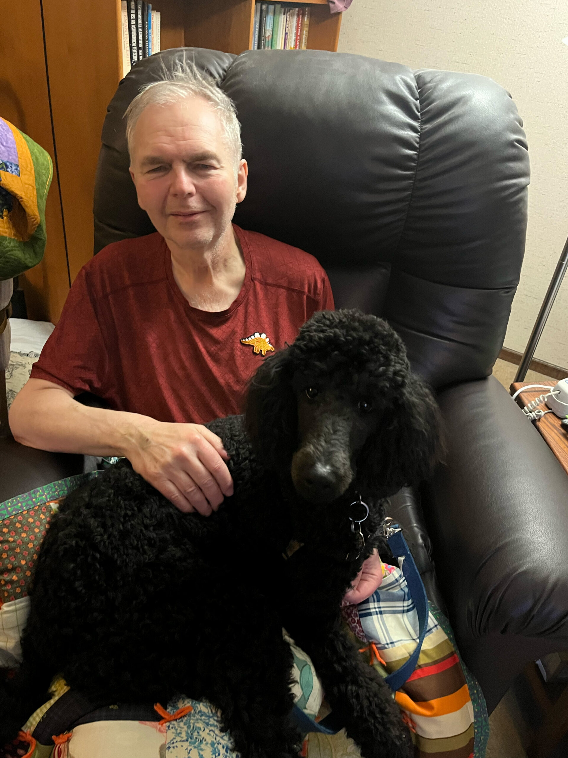Smiling older man with gray hair in a red shirt sits in a black recliner, with a black poodle on his lap and a colorful quilt nearby.